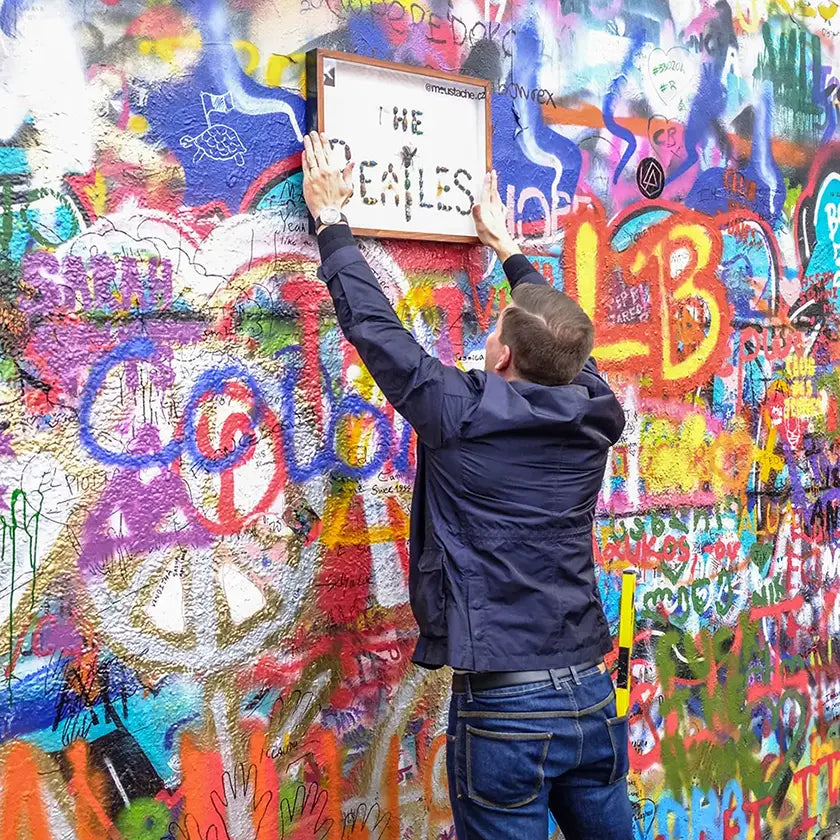 Streetartový umělec moustache připevňuje obraz s nápisem ‚The Beatles‘ vytvořený z reálných brouků na Lennonovu zeď v Praze.